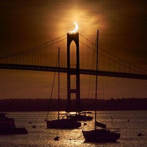 May include: A crescent moon sets behind a bridge with sailboats in the foreground. The sky is a warm orange color and the water is reflecting the light of the setting sun.