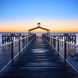 May include: A wooden pier extends out over the water towards a small gazebo with a light on top. The sky is a vibrant blue and orange, creating a beautiful sunrise scene.