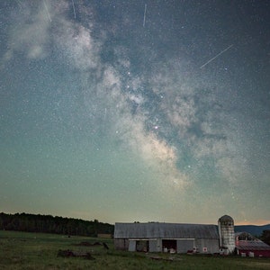 May include: A weathered barn stands in a field under a night sky filled with stars, including the Milky Way galaxy and several shooting stars.