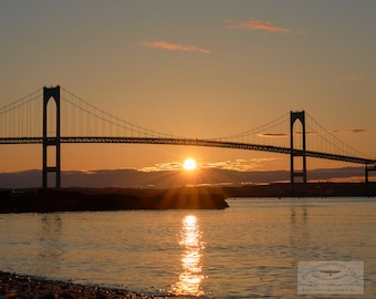 May include: A scenic photograph of a suspension bridge silhouetted against a vibrant sunset. The sun, a bright orb, is positioned behind the bridge, casting golden rays across the water. The image captures the tranquil beauty of the natural landscape.