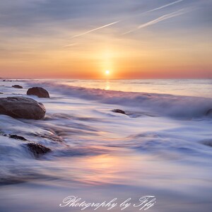 May include: A scenic photograph of a beach at sunset. The sky transitions from orange to blue, with the sun setting over the ocean. Waves crash on the shore, and large rocks are visible. The image is signed "Photography by Ffj".