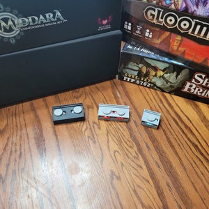 May include: Three small, rectangular game pieces. The first piece is black with two silver circles. The second piece is gray with two red circles. The third piece is gray with two blue circles.