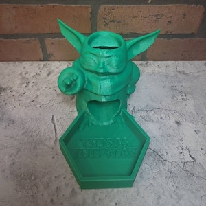 May include: Green 3D printed figurine of a character from the Star Wars franchise, known as "The Child" or "Baby Yoda", holding a small object in its hand. The figurine is standing on a hexagonal base with the words "This is the way" inscribed on it.
