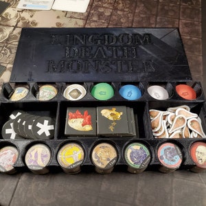 Puede incluir: Una caja organizadora negra con las palabras "Kingdom Death Monster" en relieve en la tapa. La caja está dividida en compartimentos y contiene varias piezas de juego, incluyendo fichas, cartas y miniaturas.