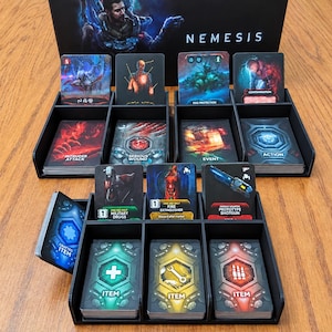 May include: A black plastic board game card organiser containing various Nemesis game cards. The organiser features multiple compartments holding decks labelled with text such as Intruder Attack, Serious Wound, Event, Action, Military Drugs, Fire Extinguisher, Prototype Shotgun, and Item.
