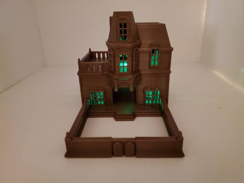 May include: A brown 3D printed model of a two-story house with a green glow coming from the windows. The house has a fence around it.