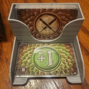 May include: A gray plastic card holder with two decks of cards. The top deck has a card with a brown background and two crossed swords. The bottom deck has a card with a green background and a white circle with a plus sign and the number one.