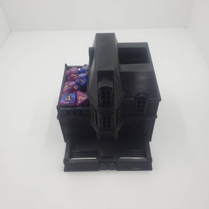 May include: A black 3D printed dice tower shaped like a house with a roof, windows, and a door. The tower has a compartment for dice and a tray for rolling dice. The tower is filled with purple and blue dice with gold numbers.