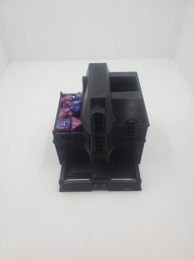 May include: A black 3D printed dice tower shaped like a house with a roof, windows, and a door. The tower has a compartment for dice and a tray for rolling dice. The tower is filled with purple and blue dice with gold numbers.