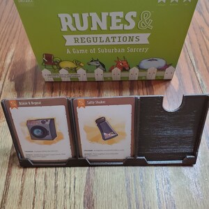 May include: A green box with the text "Runes & Regulations: A Game of Suburban Sorcery" on the front. The box is sitting on a wooden table. There are two cards on the table, one with a washing machine and the text "Rinse & Repeat" and the other with a salt shaker and the text "Salty Shaker".