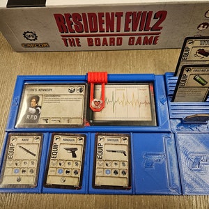 May include: A blue board game organizer with slots for cards and tokens. The organizer holds cards labeled "Knife", "Handgun", and "Shotgun". The game box, "Resident Evil 2 The Board Game", is visible in the background.