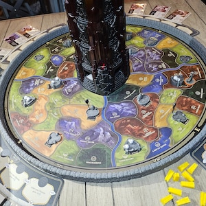 May include: A game board with a circular map featuring various coloured regions and a central tower. The tower is dark brown and has a red light at the top. There are small white figures on the board and yellow plastic pieces on the table.