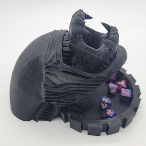 May include: A black 3D printed dice tower shaped like an alien head with a large open mouth. The tower has a circular base with a gear design. There are several multi-coloured dice with gold numbers inside the tower and on the base.