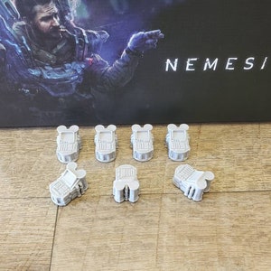Puede incluir: Siete piezas de juego de plástico gris con bordes redondeados y un diseño en relieve. Las piezas tienen forma de pequeños robots con dos orejas en la parte superior. Las piezas están dispuestas en semicírculo sobre una superficie de madera. La palabra "NEMESIS" es visible en el fondo.