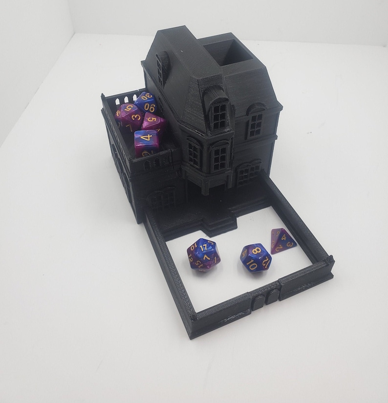 May include: A black 3D printed dice tower shaped like a house with a removable tray. The tower has a roof with a small opening for dice to fall through. The tray is open on one side and has a black border. There are several blue and purple dice with gold numbers on the tray and inside the tower.