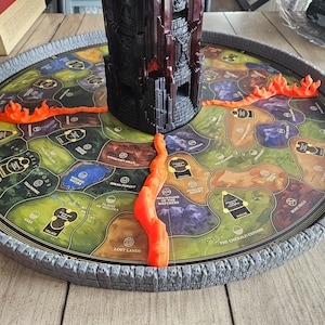 May include: A circular board game with a grey stone wall border. The board features a map with various locations, including "The Emerald Expanse" and "Lost Lands". Orange flames surround the board.