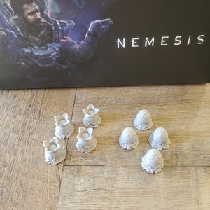 May include: Eight white plastic game pieces shaped like alien eggs. The pieces are arranged in a semi-circle on a wooden surface. The game box for the board game "Nemesis" is visible in the background.