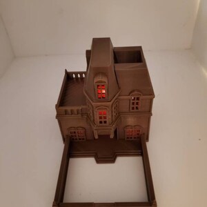 May include: A brown 3D printed model of a house with a red lit window. The house has a balcony and a small yard.