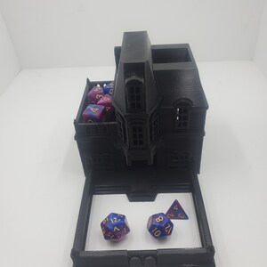 May include: A black 3D printed house shaped dice tower with a removable tray. The tower has a small roof with a dormer window. The tray has three dice, two are blue and purple with gold numbers, and one is a purple and blue triangle with gold numbers.