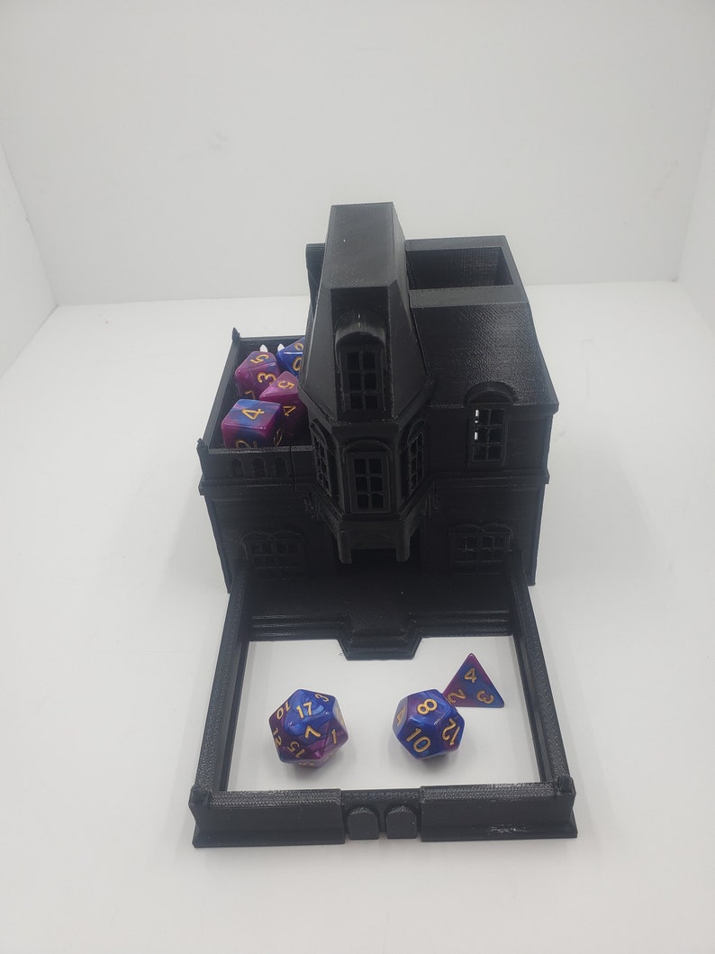 May include: A black 3D printed house shaped dice tower with a removable tray. The tower has a small roof with a dormer window. The tray has three dice, two are blue and purple with gold numbers, and one is a purple and blue triangle with gold numbers.