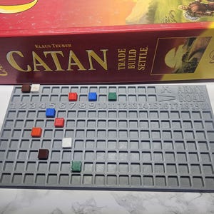 May include: A Catan board game set with a gray game board and colorful game pieces. The game board has numbered squares and the text "Army Road". The Catan box is red and yellow.