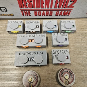 May include: A set of gray plastic ammo counters for the Resident Evil 2 board game. Each counter has a different weapon name and ammo capacity printed on it. The counters are arranged in two rows on a wooden surface.