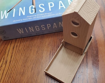 May include: A beige wooden birdhouse with two round openings and a tray base. The birdhouse is sitting on a wooden table next to a board game box with the title "Wingspan" on it. The box is blue with a white bird illustration.