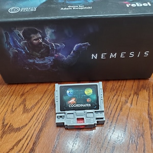 May include: A gray box with the words "Awaken Realms" and "Nemesis" on it. The box is sitting on a wooden surface. A small gray device with a screen that says "Coordinates" is sitting on the box.
