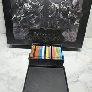 Pode incluir: Um organizador preto com tampa para guardar cartas. O organizador tem uma etiqueta que diz "Return to Dark Tower". O organizador tem vários compartimentos para guardar cartas de diferentes cores.