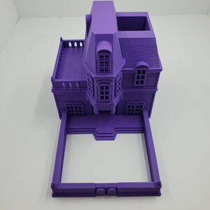 May include: A purple 3D printed model of a house with a square base and a gabled roof. The house has a balcony, windows, and a door. The model is made of plastic and is about 6 inches tall.