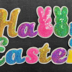 May include: Happy Easter is spelled out in colorful sequins with two pink and green bunny figures.