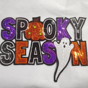 Op de afbeelding: Een met pailletten versierde patch met Halloween-thema met de woorden "Spooky Season". De letters zijn in verschillende kleuren, waaronder paars, oranje en zwart. Een pompoen en een spook zijn ook in het ontwerp opgenomen.