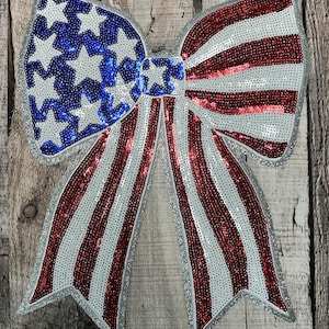 May include: A large, red, white, and blue sequined bow with a star-spangled design. The bow is made of shiny sequins and has a silver outline.