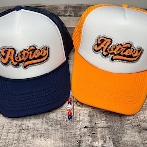 May include: Two white and orange trucker hats with a sparkly orange "Astros" logo on the front. The hats have a mesh back and a navy blue brim.