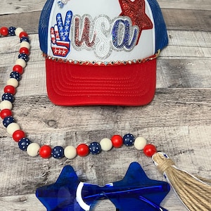 May include: A red, white, and blue trucker hat with a sparkly "USA" design and a peace sign with stars and stripes. The hat is paired with a red, white, and blue beaded necklace and blue star-shaped sunglasses.