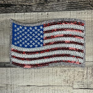 May include: A sequined American flag patch with a silver glitter border. The flag features a blue field with white stars and red and white stripes. The patch is rectangular and has a wavy top edge.