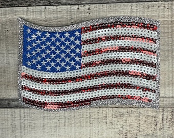 May include: A sequined American flag patch with a silver glitter border. The flag features a blue field with white stars and red and white stripes. The patch is rectangular and has a wavy top edge.