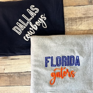 Puede incluir: Dos mantas dobladas. Una es azul marino con "DALLAS cowboys" bordado en blanco. La otra es gris con "FLORIDA gators" bordado en azul y naranja. Las mantas están sobre una superficie de madera.