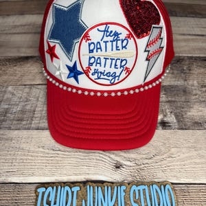 Puede incluir: Gorra de béisbol roja y blanca con la parte trasera de malla. La gorra presenta una estrella azul, un corazón rojo con lentejuelas y un rayo. La parte delantera de la gorra tiene el texto "Hey Batter Batter Swing!" y está adornada con perlas.