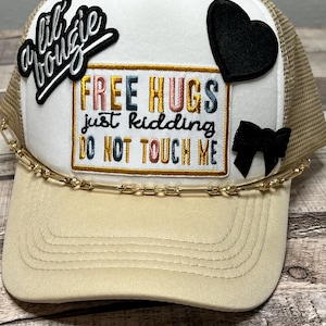 May include: A white and beige trucker hat with a gold chain trim. The hat has several patches, including a black heart, a black and white bow, and a black patch that says "a lil bougie". The hat also has an embroidered patch that says "FREE HUGS just kidding DO NOT TOUCH ME".