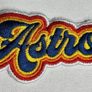 May include: A colorful embroidered patch with the word "Astros" in a retro font. The letters are blue with yellow and orange outlines.