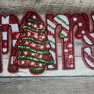 Puede incluir: Decoración navideña con lentejuelas que deletrea la palabra "MARY". La "M" es a cuadros rojos y blancos, la "A" presenta un árbol de Navidad y la "RY" diseños de bastones de caramelo. Los colores son rojo, verde y blanco.