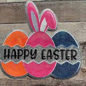 Könnte beinhalten: Ein glitzerndes Frohe Ostern Schild mit drei Eiern, ein pinkfarbenes mit Hasenohren, ein orangefarbenes und ein blaues.