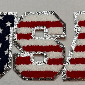 May include: USA spelled out in large, silver-trimmed letters. Each letter is made of red and white striped fabric, resembling the American flag. The "U" and "S" are blue with white stars.