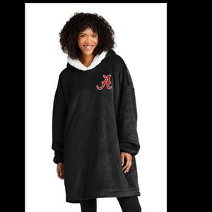 May include: Black oversized hooded blanket with a white fleece-lined hood. The blanket features a red embroidered letter "A" on the chest. The model is wearing black leggings.