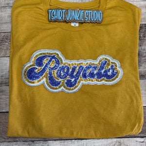 May include: Mustard yellow t-shirt with the word "Royals" in a blue and silver sequined design. The shirt also features the text "Tshirt Junkie Studio" in a blue font. The shirt is folded and laid flat on a wooden surface.