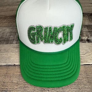 Op de afbeelding: Groene en witte truckerpet met het woord "GRINCHY" in groene glitterletters op de voorkant. De pet heeft een groene klep en mesh achterkant, met een wit voorpaneel. Klassieke truckerpet stijl.