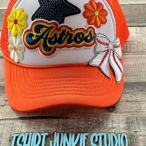 May include: Orange and white trucker hat with a white patch that says "Astros" in a retro font. The patch has a black star with sequins, a yellow daisy, and an orange daisy. There is also a white bow with red stripes and a baseball stitch design.