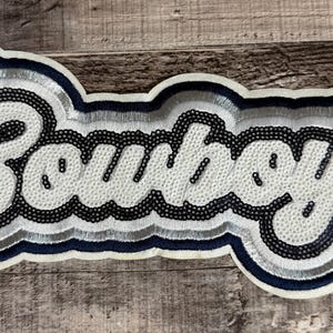 May include: A white and navy blue embroidered patch with the word "Cowboys" in a script font. The patch has a layered design with silver and black accents, and a navy blue border. The patch is on a wooden surface.