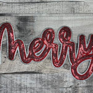May include: A decorative sign spelling "Merry" in a cursive font. The letters are red with a sequined texture, outlined in silver glitter. The sign is set against a weathered, gray wooden background.